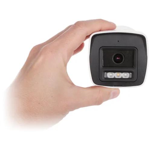 Osoba trzyma w dłoni kamerę monitoringu o prostokątnym kształcie z widocznym obiektywem i diodami LED. Kamera ma biało-czarną obudowę i jest sfotografowana na białym tle.