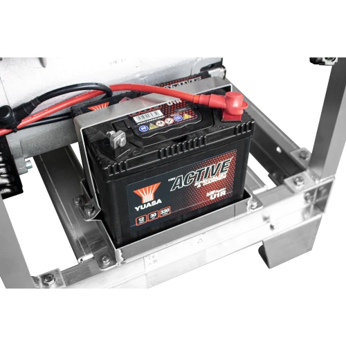 Zdjęcie przedstawia akumulator Yuasa Active umieszczony w metalowej ramie agregatu prądotwórczego. Widać czerwone i czarne przewody podłączone do biegunów akumulatora.