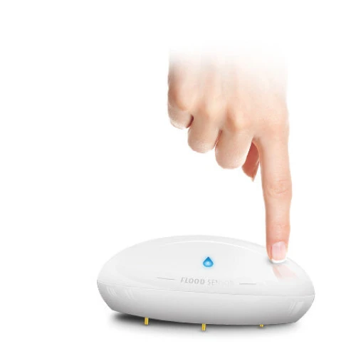 Na zdjęciu znajduje się biały czujnik zalania wodą marki Fibaro z niebieskim wskaźnikiem na górze. Obok czujnika widoczny jest palec wskazujący, co sugeruje jego niewielki rozmiar i łatwość obsługi.