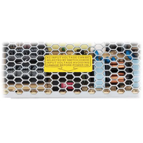 Zdjęcie przedstawia zbliżenie na metalową kratkę ochronną z widocznymi elementami elektronicznymi wewnątrz, w tym kondensatory i cewki. Na kratce znajduje się żółta naklejka z ostrzeżeniem dotyczącym wyboru napięcia wejściowego.