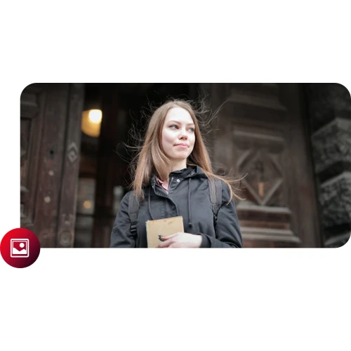 Na zdjęciu znajduje się młoda kobieta z długimi włosami, ubrana w ciemną kurtkę, trzymająca książkę i stojąca przed drewnianymi drzwiami. W tle widać detale architektoniczne budynku.