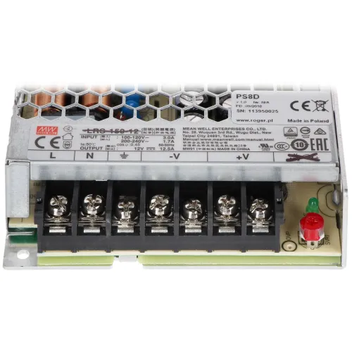 Zdjęcie przedstawia zasilacz impulsowy z widocznymi złączami śrubowymi i etykietą informacyjną na obudowie. W tle widać metalową siatkę ochronną oraz elementy elektroniczne.